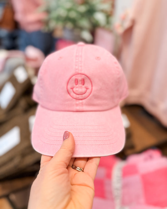 Pink cap with smiley face design held by a hand against a blurred indoor background