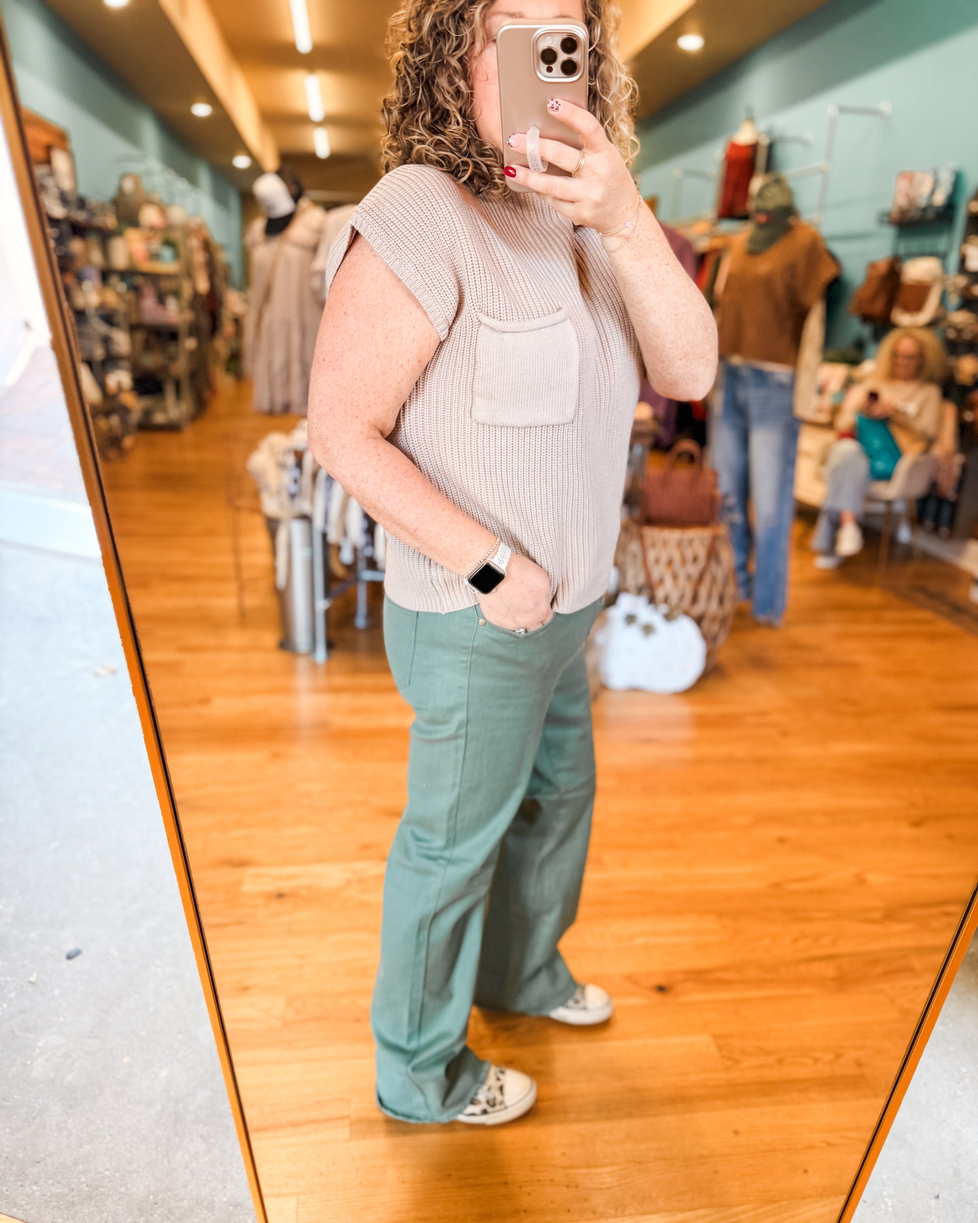 Person taking a mirror selfie wearing a sleeveless top and green pants in a store.