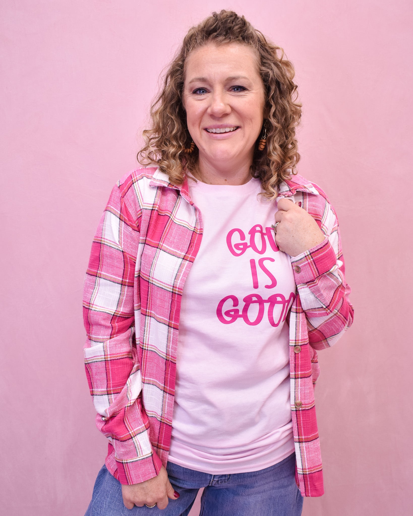 Person wearing a pink plaid shirt over a white t-shirt with 'God is Good' text on a pink background