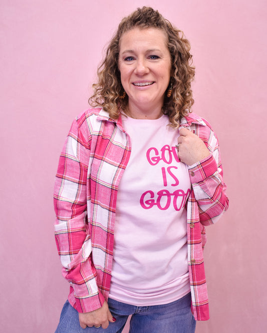 Person wearing a pink plaid shirt over a white t-shirt with 'God is Good' text on a pink background