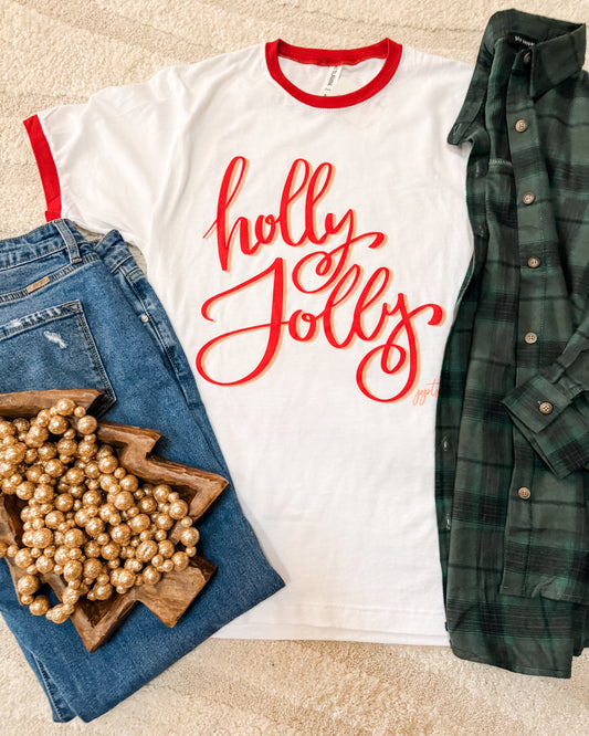 White t-shirt with 'holly jolly' text, blue jeans, plaid shirt, and decorative items on a beige surface.