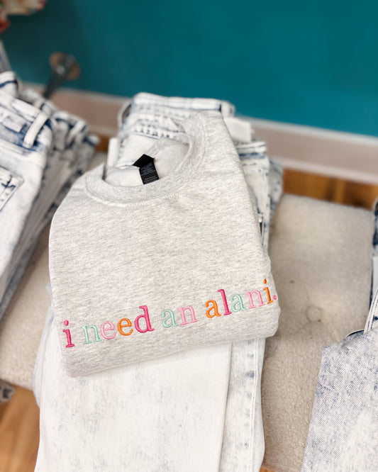 Gray sweatshirt with colorful text 'i need an alani' on a rack.