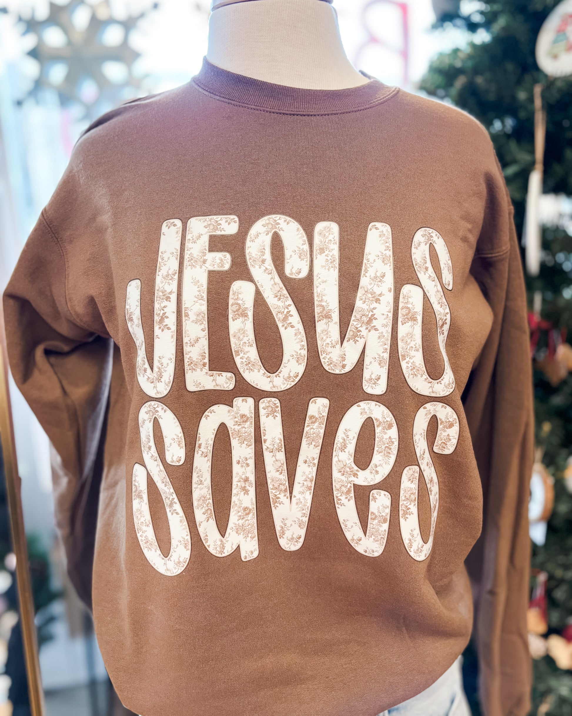 Brown sweatshirt with 'Jesus Saves' text on a mannequin against a blurred indoor background.