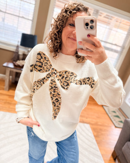 Person wearing a white sweater with a large leopard print bow, taking a mirror selfie.