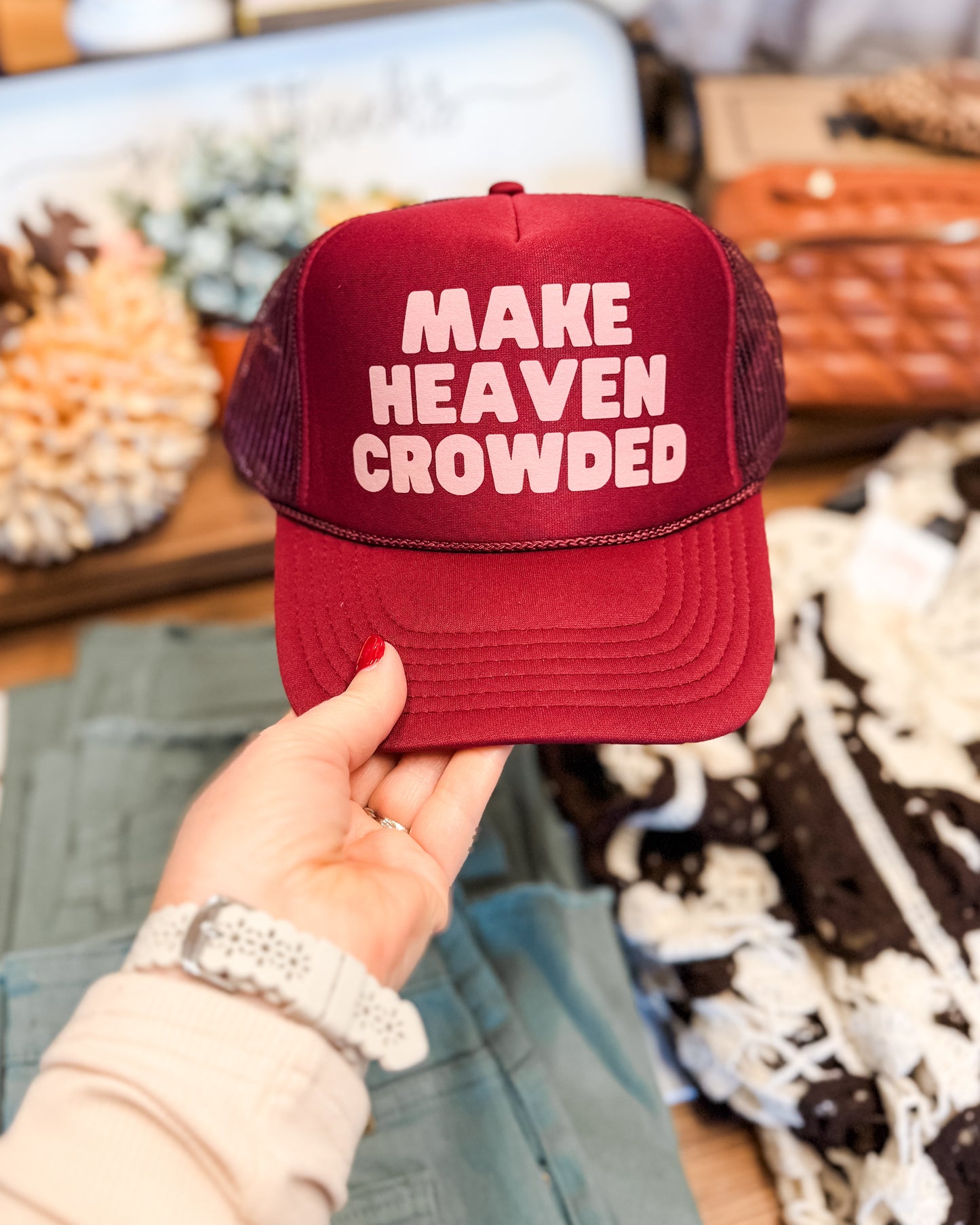 Red cap with 'MAKE HEAVEN CROWDED' text held by a person in a store setting.