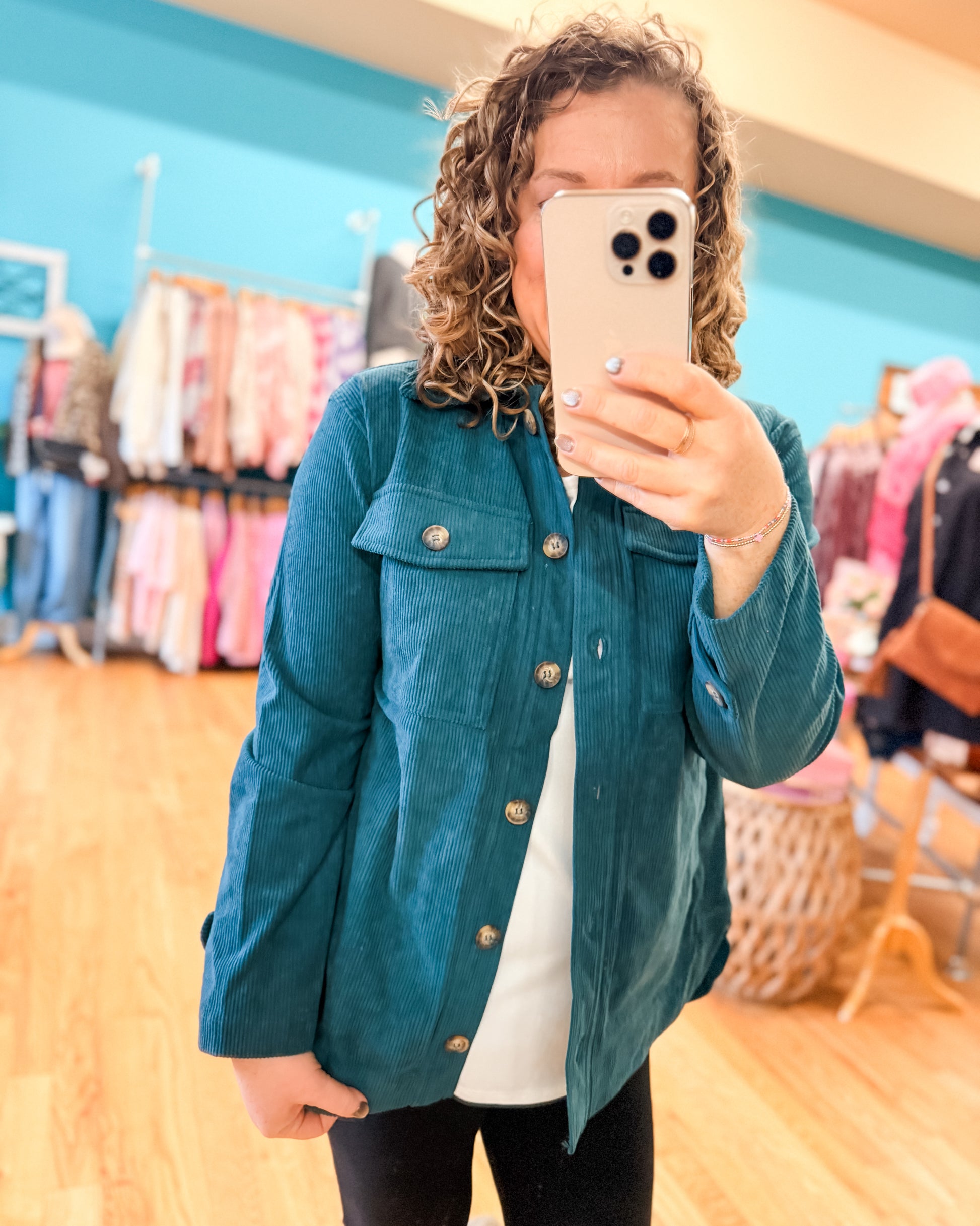 Person wearing a teal jacket taking a mirror selfie in a clothing store.