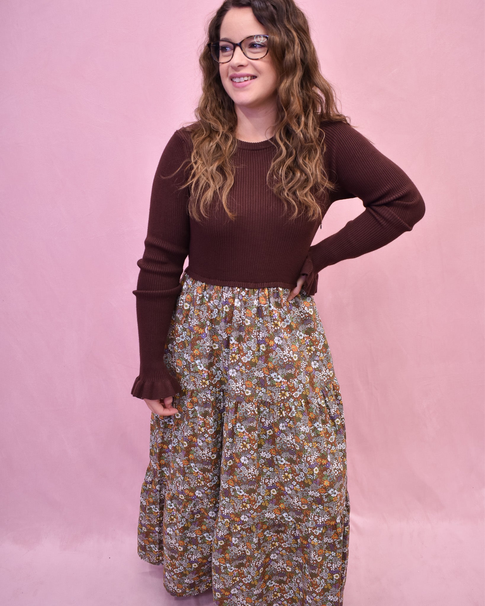 Woman wearing a brown sweater and floral pants against a pink background