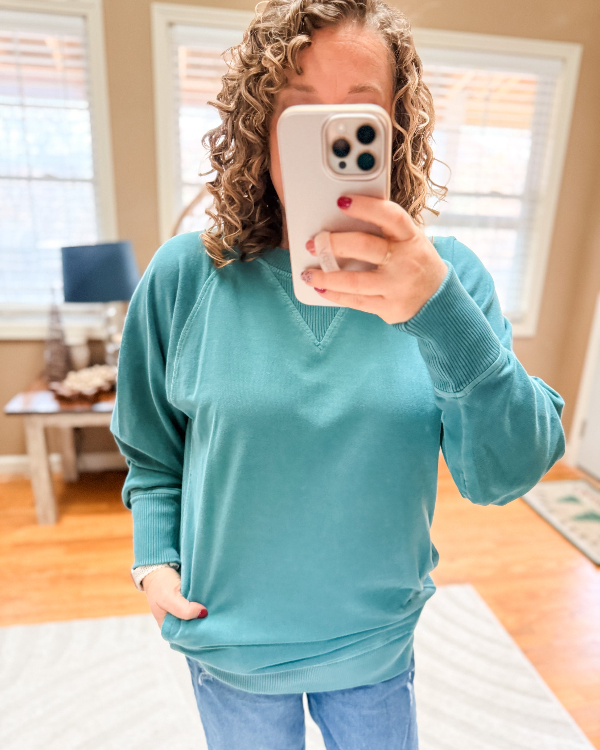 Person wearing a teal sweater taking a mirror selfie in a room with wooden floors and a window.