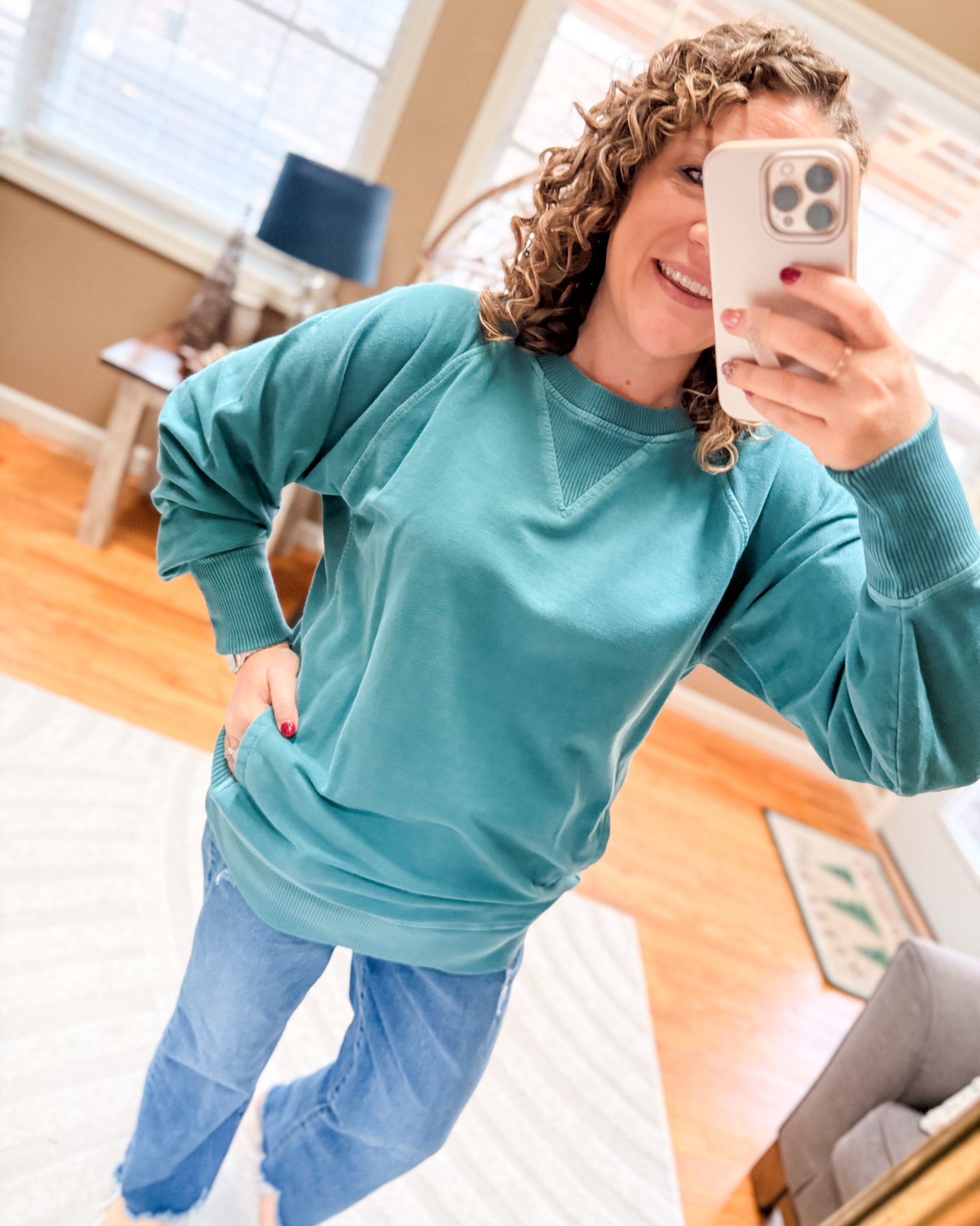 Woman in teal sweatshirt and blue pants taking a mirror selfie in a home setting.