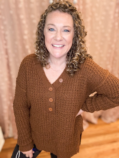 A smiling woman wearing a brown V-neck sweater with button trim details.
