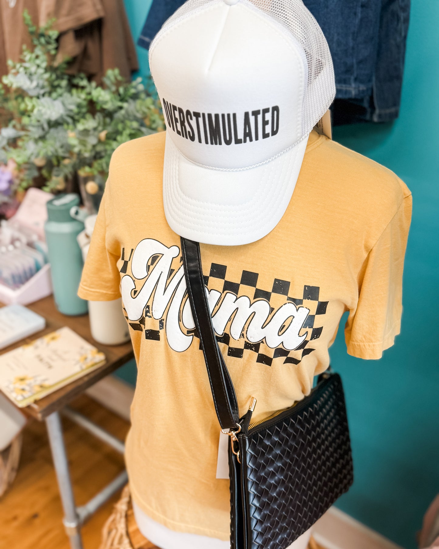 Mannequin wearing a yellow t-shirt with 'Mama' print, white cap with 'OVERSTIMULATED' text, and black handbag.