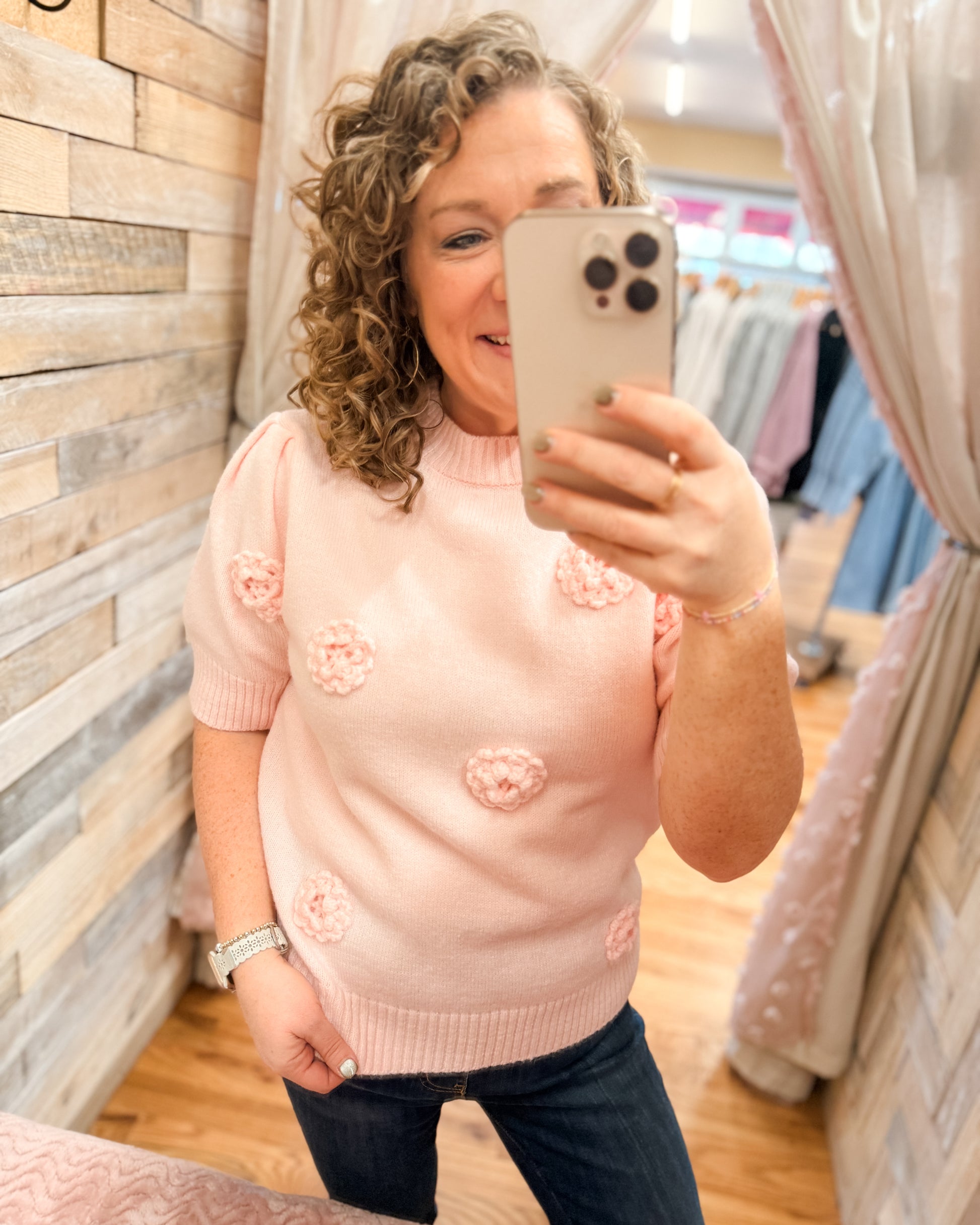 Woman taking a mirror selfie wearing a pink sweater with floral patterns in a store.