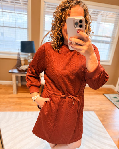 Person wearing a rust-colored dress taking a mirror selfie in a room with wooden flooring and a lamp.