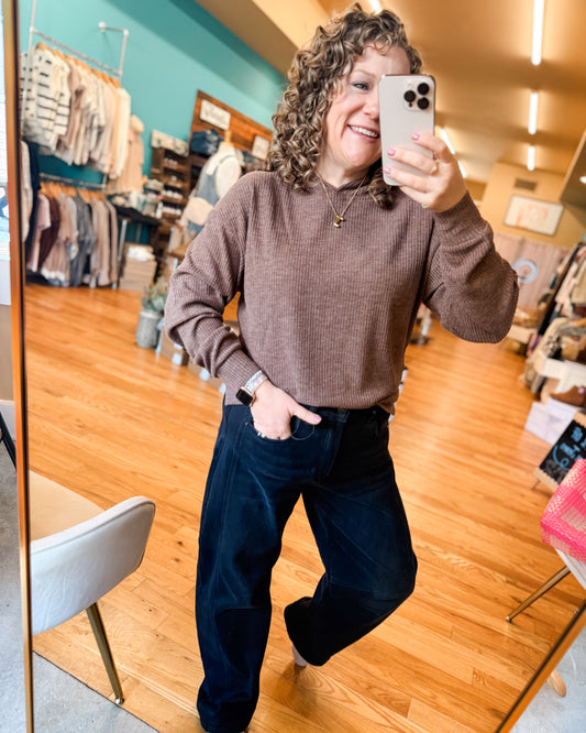 Person taking a mirror selfie in a clothing store wearing a brown sweater and dark pants.