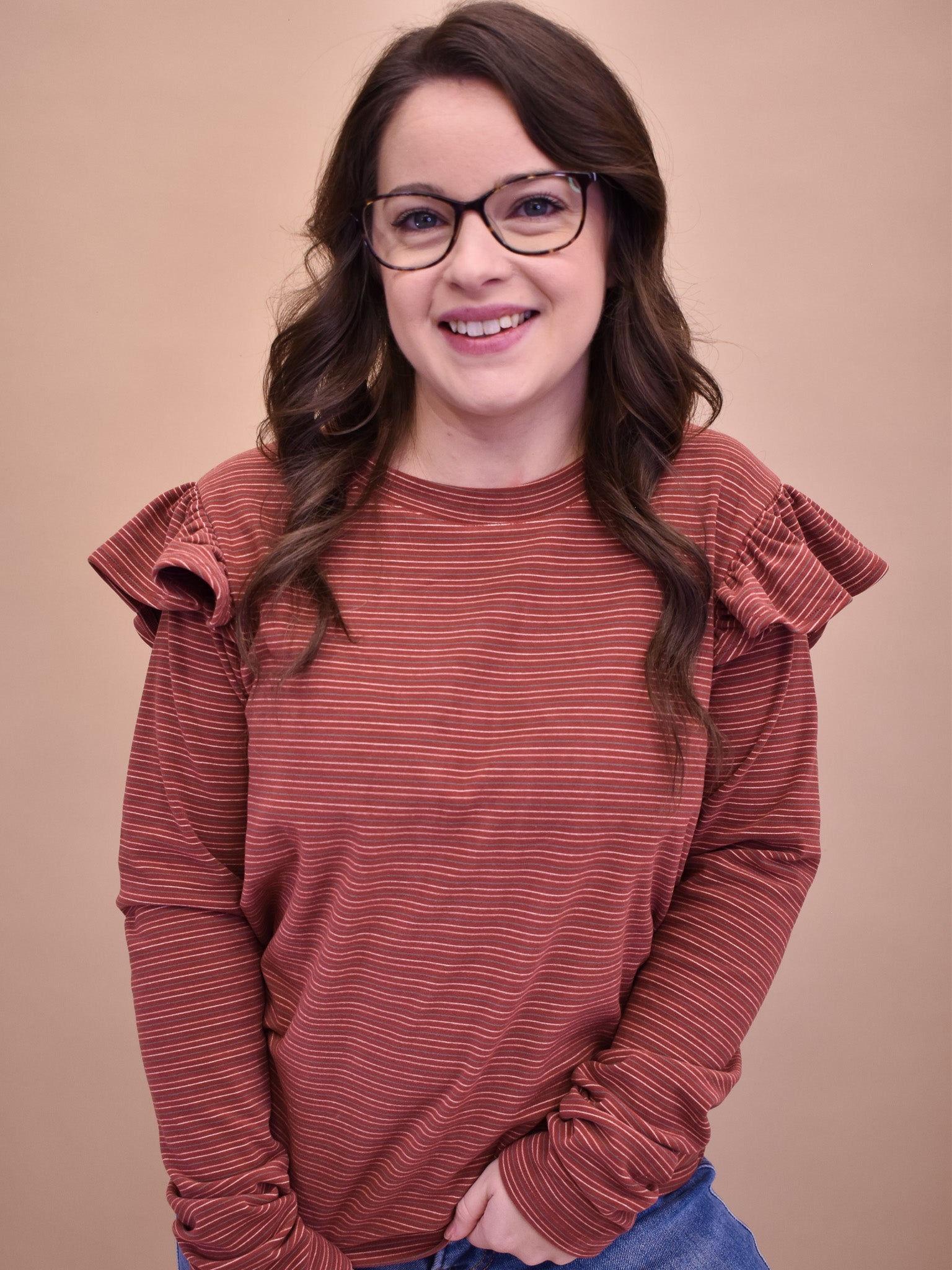 A person wearing a brown, long sleeve, ruffle shoulder top with glasses, smiling against a beige background.