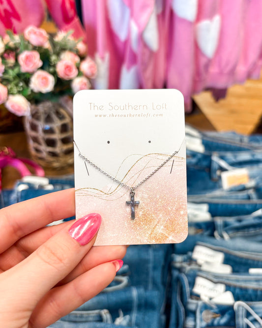 Necklace with a cross pendant on a card held by a hand, with jeans and flowers in the background