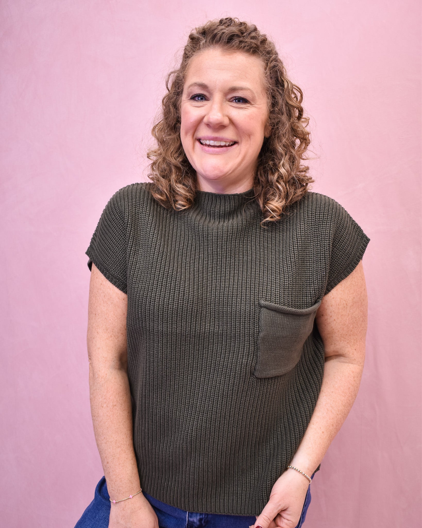 Woman wearing a green ribbed knit sleeveless sweater against a pink background