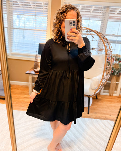Person wearing a black dress taking a mirror selfie in a room with a couch and window blinds.
