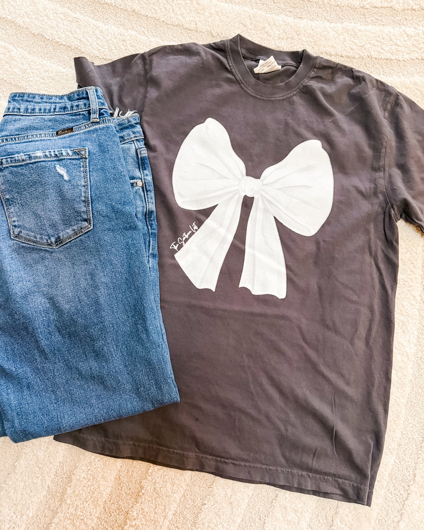 Gray t-shirt with a white bow graphic and blue jeans on a beige surface