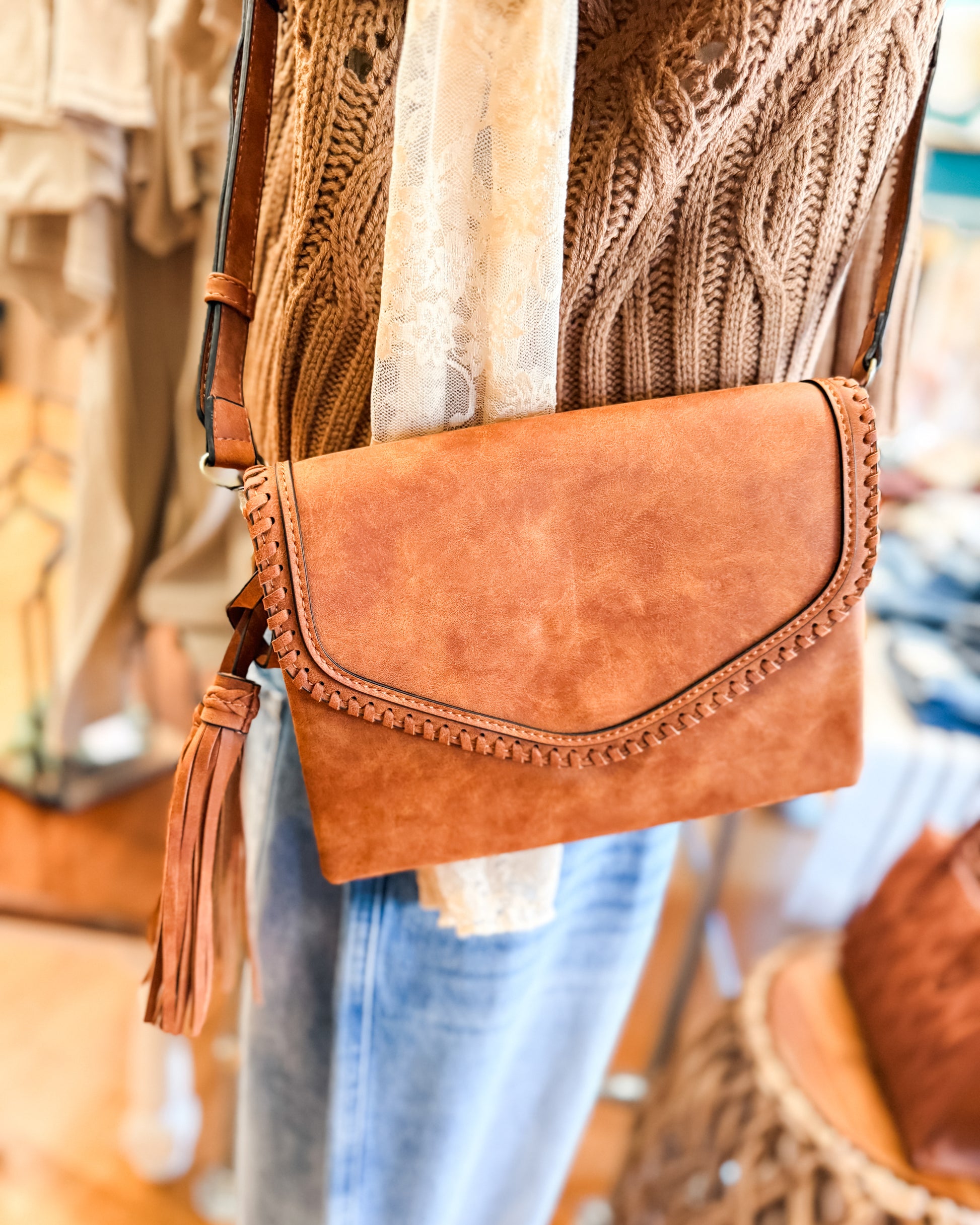 Brown leather clutch bag with tassel detail worn by a person in a blurred indoor setting