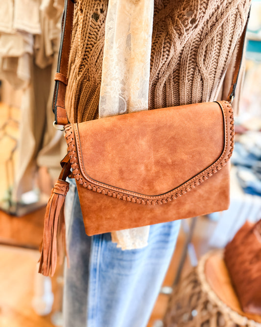 Brown leather clutch bag with tassel detail worn by a person in a blurred indoor setting