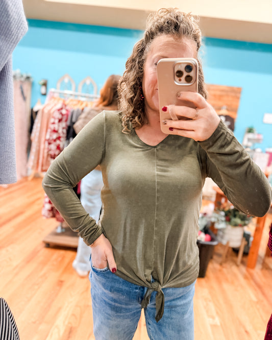 Person taking a mirror selfie wearing an olive green long-sleeve top and blue jeans in a store.
