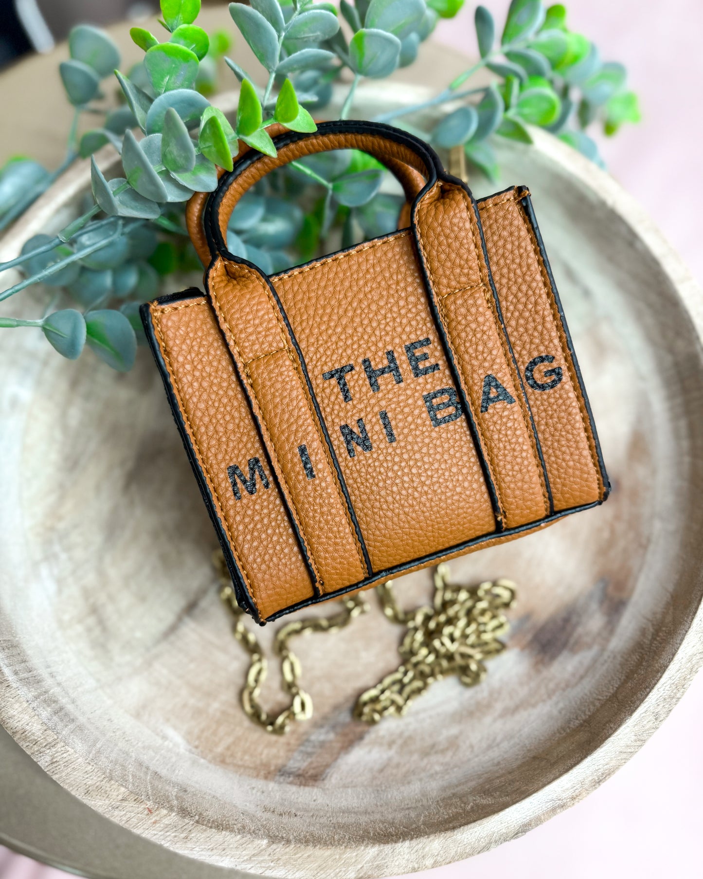 Brown leather bag charm with 'THE MINI BAG' text on a wooden surface with greenery.