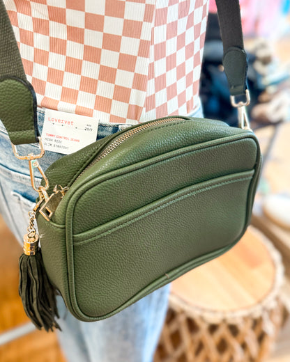 Green crossbody bag with a tassel detail worn by a person, blurred background