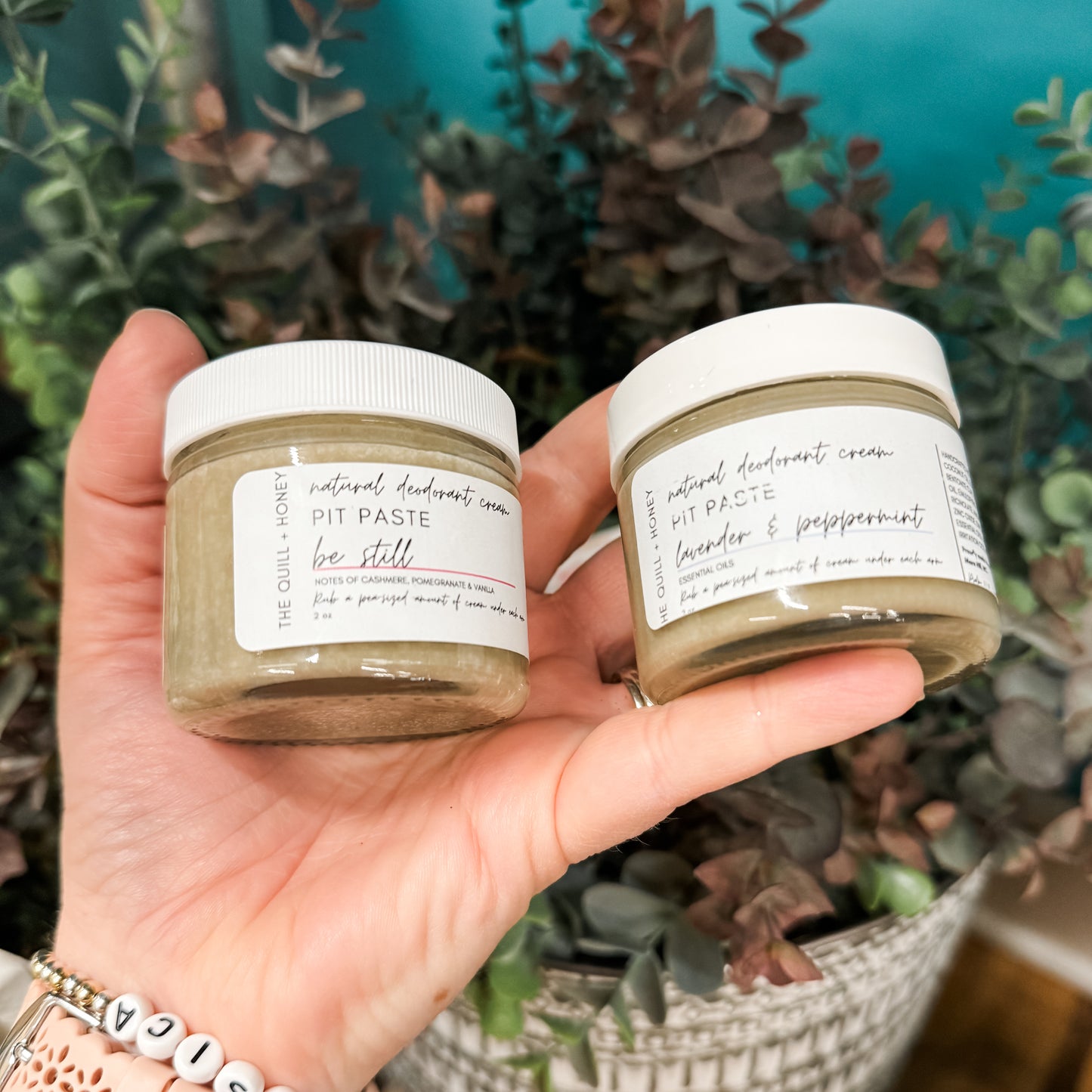 Two jars of The Quill & Honey Pit Paste, one held in hand with a plant background and the other on a shelf with a wristwatch visible, indicating a cream-colored deodorant product.