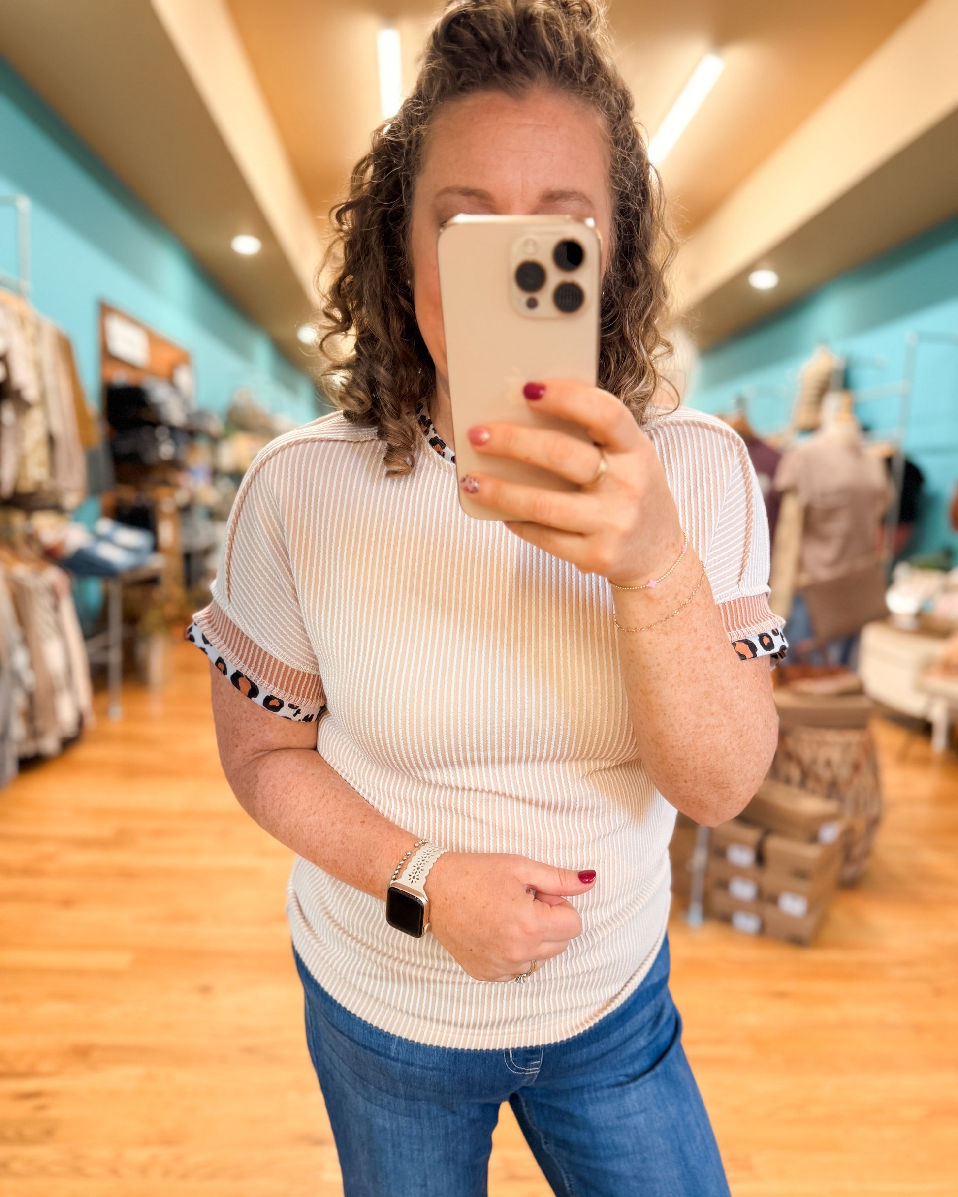 Woman taking a mirror selfie in a store wearing a white top with black and red accents and blue jeans.