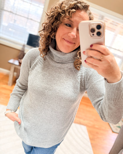 Woman in a gray sweater taking a selfie in a room with wooden flooring.