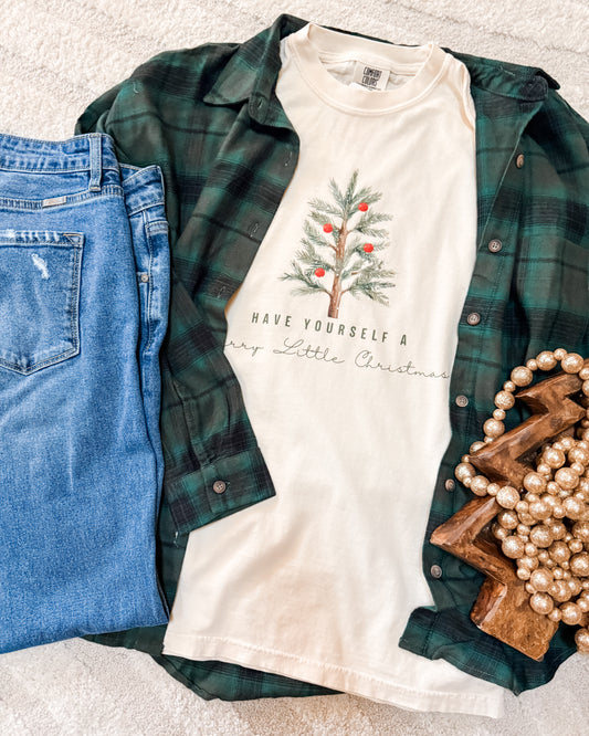 White t-shirt with Christmas tree design and text, green plaid shirt, blue jeans, and decorative items on a light gray surface.